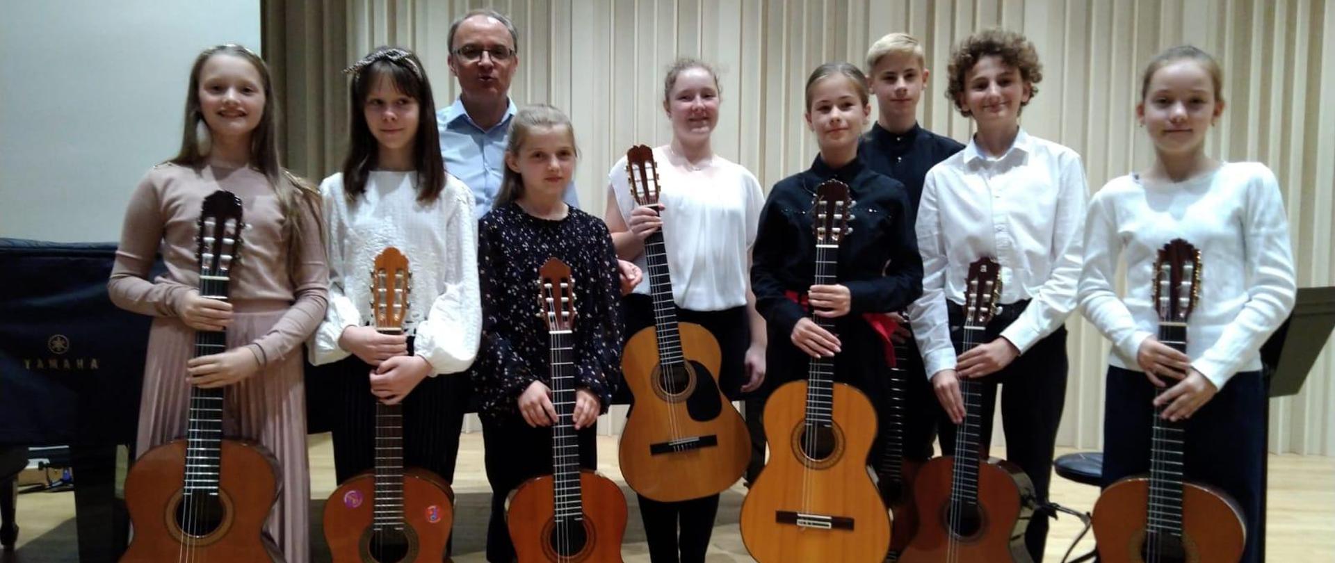 Na zdjęciu grupa uczniów. Wszyscy odświętnie ubrani. Każdy z nich trzyma gitarę. Tło obrazka w odcieniach brązu.