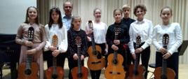 Na zdjęciu grupa uczniów. Wszyscy odświętnie ubrani. Każdy z nich trzyma gitarę. Tło obrazka w odcieniach brązu.
