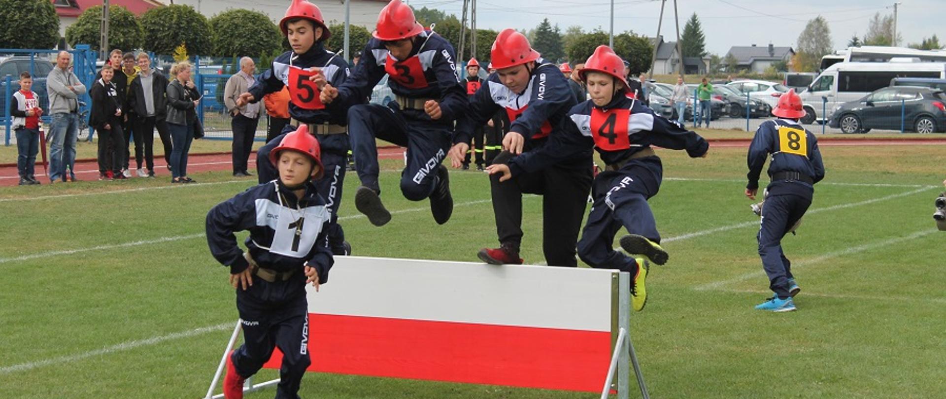 Fotografia wykonana podczas „XI Powiatowych Zawodów Sportowo-Pożarnicze Młodzieżowych Drużyn Pożarniczych Ochotniczych Straży Pożarnych na stadionie w Przecławiu. Na zdjęciu zawodnicy podczas konkurencji „Rozwinięcia bojowego”. Pięciu zawodników jednocześnie pokonuje przeszkodę w postaci ścianki. Na głowach mają ubrane hełmy strażackie koloru czerwonego.