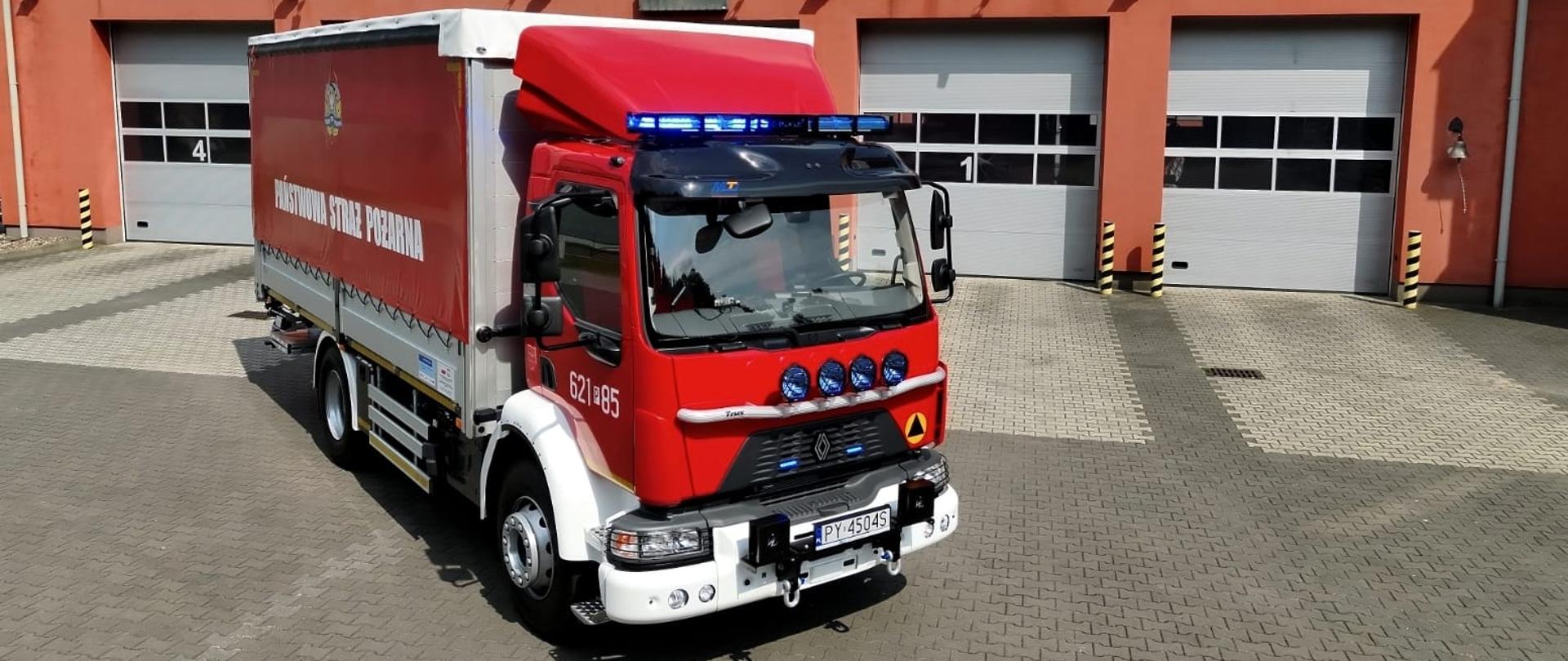 Zdjęcie przedstawia czerwono-biały samochód ciężarowy Państwowej Straży Pożarnej marki Renault, który stoi na placu z szarej kostki brukowej przed nowoczesną remizą. Pojazd ma włączone niebieskie sygnały błyskowe, numery operacyjne 621[P]85 oraz tablicę rejestracyjną PY 45045, a na jego skrzyni ładunkowej z plandeką widnieje napis „Państwowa Straż Pożarna”. W tle znajduje się pomarańczowy budynek Komendy z trzema zamkniętymi bramami wjazdowymi, nad którymi umieszczono duży, biały napis „Państwowa Straż Pożarna 998”. Całość sceny dopełnia czyste, jasnoniebieskie niebo.
