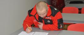 Egzamin KDR. Świetlica w Komendzie Powiatowej PSP w Rawiczu. Za stołem siedzi jeden z egzaminowanych druhów OSP. Jest pochylony nad testem. W prawej ręce trzyma długopis. Ma na sobie czerwony polar z napisem STRAŻ.