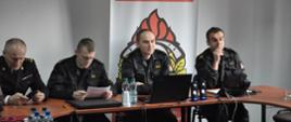 Na pierwszym planie komendanci KP PSP w Ostrowcu oraz oficerowie podsumowujący ćwiczenia w sali konferencyjnej. W tle logo KP PSP w Ostrowcu Św.