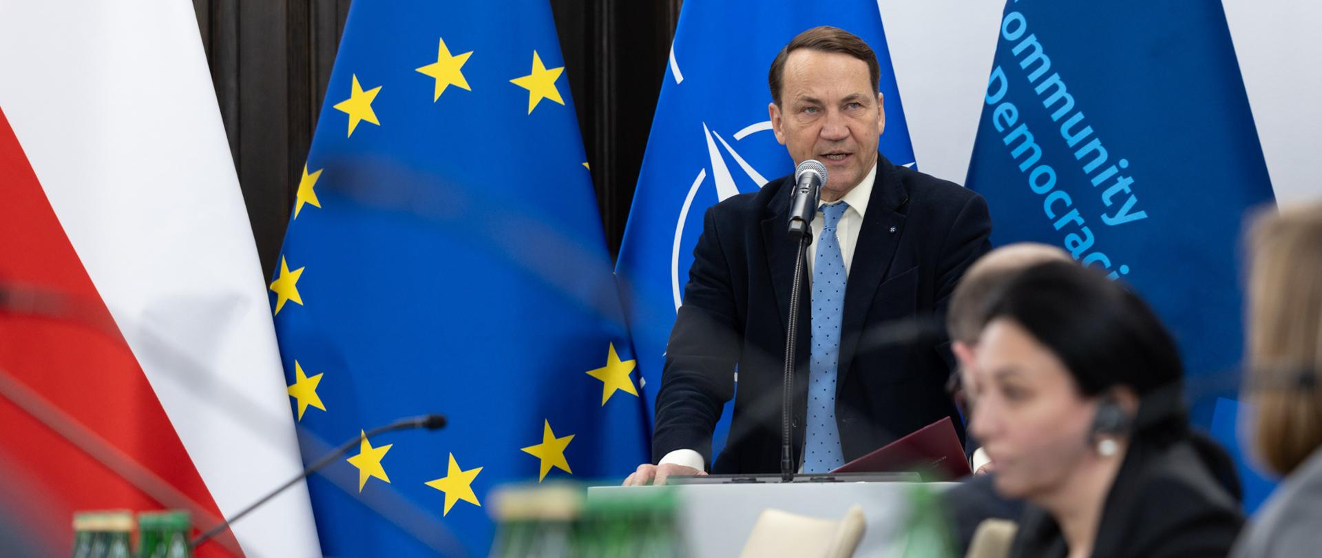 Mężczyzna przemawia przy mównicy podczas oficjalnego spotkania, na tle flag Polski, Unii Europejskiej, NATO. Na pierwszym planie widoczni są uczestnicy siedzący przy stole konferencyjnym.