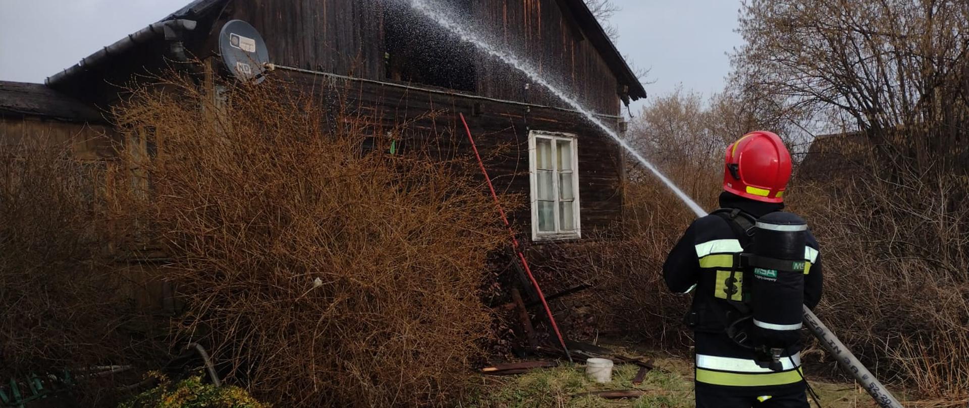 Na zdjęciu stoi strażak ubrany w aparat ochrony dróg oddechowych, który trzyma prądownicę. Jest on odwrócony oraz gasi tlące się elementy konstrukcji dachu budynku, który objęty był pożarem.
