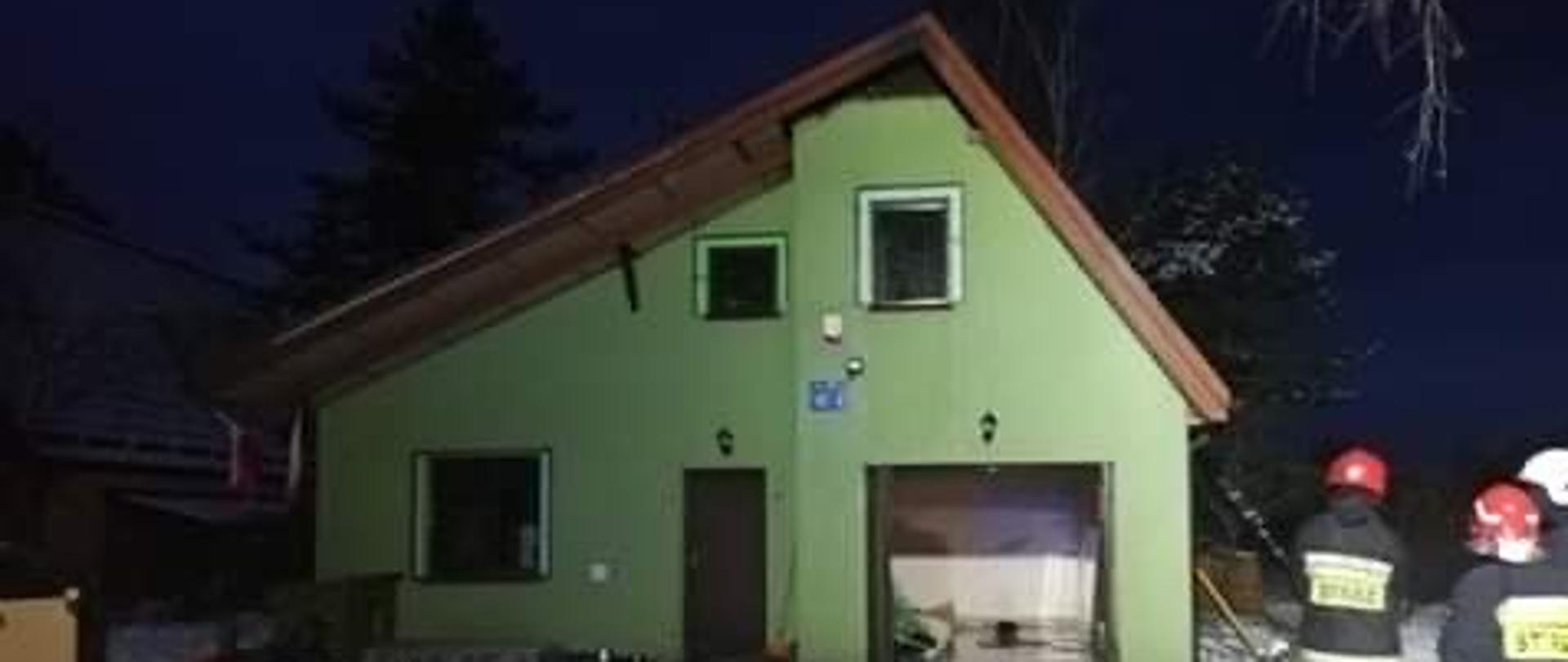 Na zdjęciu widać budynek mieszkalny jednorodzinny, z elewacją w kolorze zielonym, pokryty blachą oraz podbitką dachową, która w górnej części została nadpalona, następnie ugaszona i częściowo rozebrana.
W budynku widoczne są trzy okna plastikowe i jedne metalowe drzwi wejściowe nad nimi oświetlenie zewnętrzne oraz otwarte metalowe, drzwi garażowe nad nimi oświetlenie zewnętrzne budynku. Na środku budynku umiejscowiona jest czujka od alarmu oraz biały numer na niebieskim tle. Przed domem jest utwardzony plac manewrowy wykonany z kostki brukowej o kolorze bordowym, częściowo pokryty śniegiem na którym rozwinięty został odcinek linii gaśniczej. Z lewej strony budynku widać dwie flagi biało-czerwone oraz elementy wystroju budynku, które zostały wyniesione z wewnątrz mieszkania. Z prawej strony widać trzech strażaków, ubranych w ubranie specjalne w kolorze granatowym oraz w dwa hełmy czerwone i jeden biały. Z tej samej strony widać także drabinę strażacką, opartą o dach budynku.