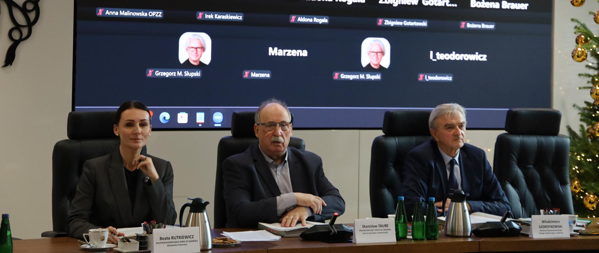 posiedzenie rady, przy stole siedzą trzy osoby, za nimi na telebimie inni członkowie posiedzenia