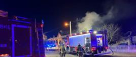 Na zdjęciu znajdują się samochody pożarnicze, strażacy oraz w tle dym z dogaszanego budynku mieszkalnego