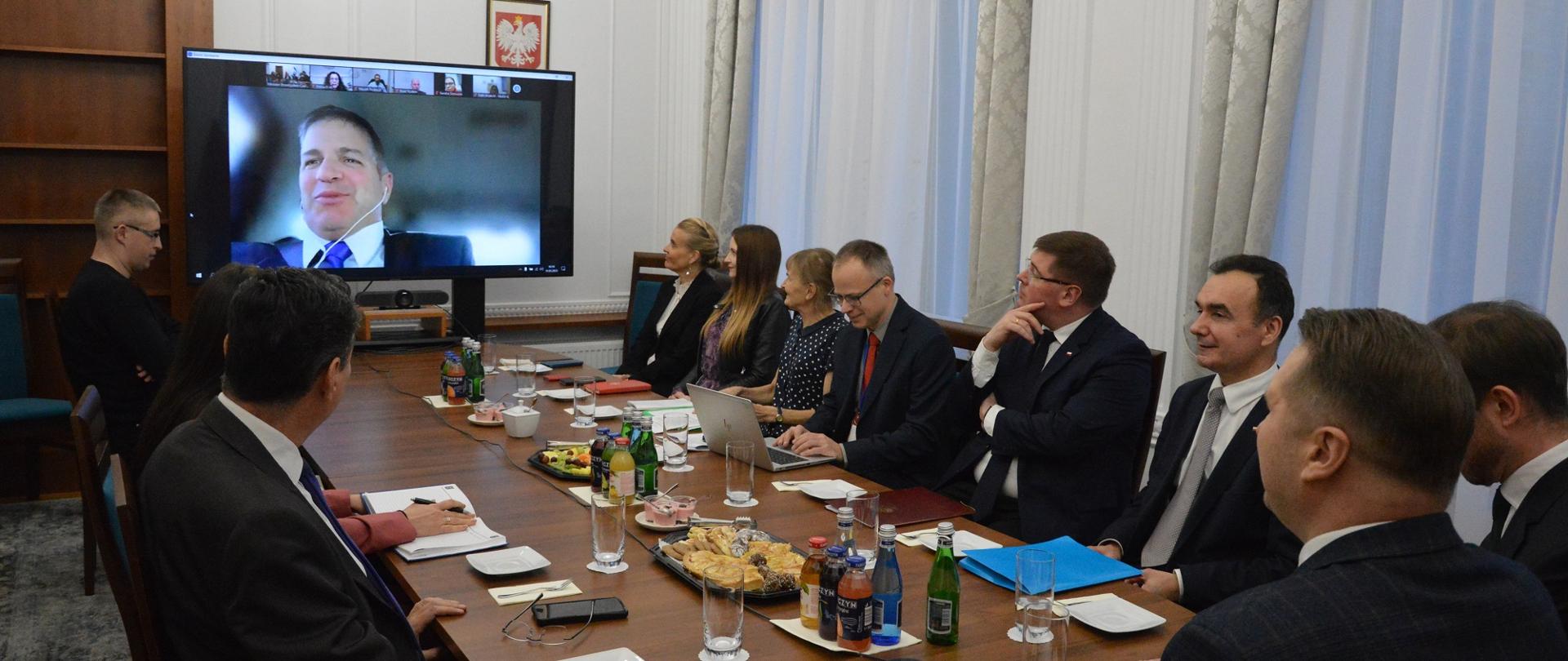 Przy podłużnym stole siedzi grupa ludzi, patrzą na znajdujący się nad jednym z końców stołu duży monitor na którym widać twarz mężczyzny. Z lewej biała ściana z dwoma oknami.