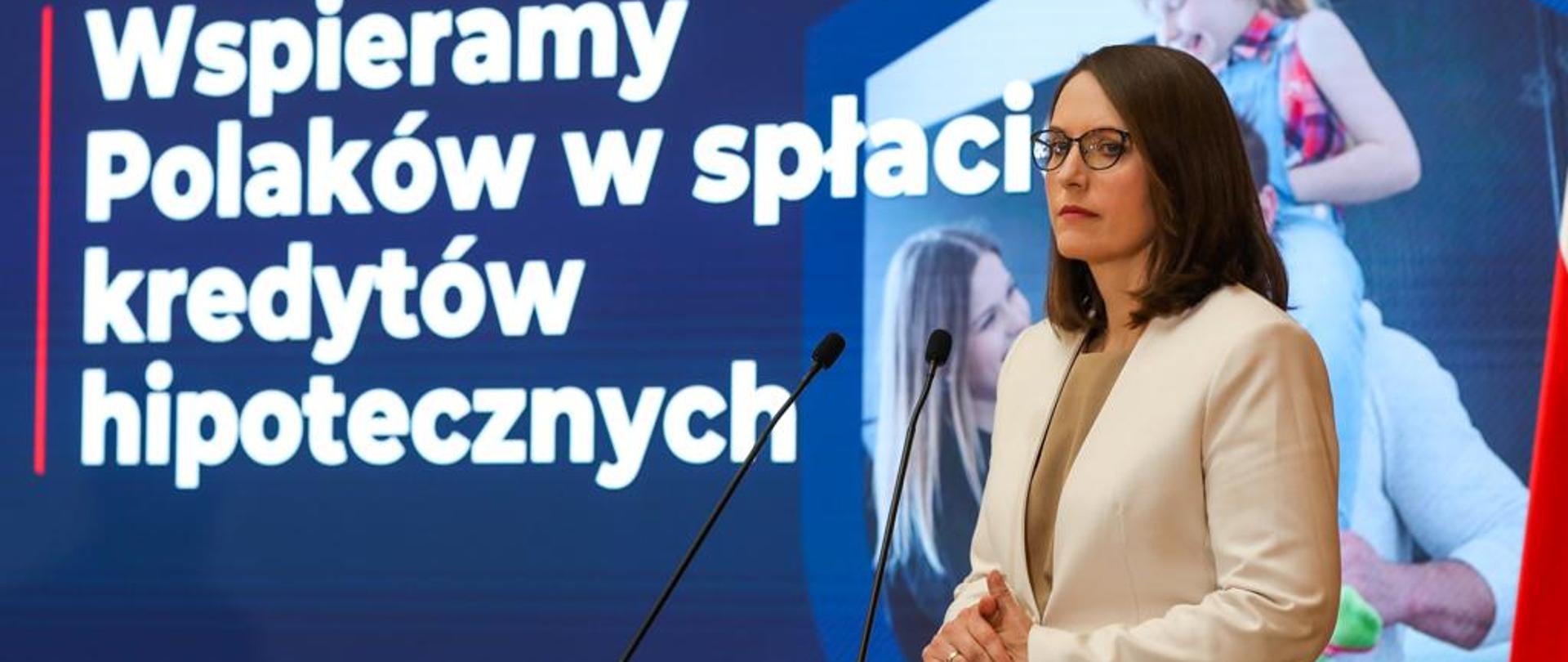 Minister finansów Magdalena Rzeczkowska podczas wystąpienia. W tle napis Tarcza Antyputinowska. Wspieramy Polaków w spłacie kredytów hipotecznych.