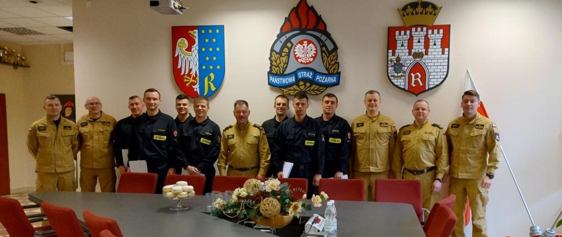 Zdjęcie wykonane w jednym z pomieszczeń komendy miejskiej Państwowej Straży Pożarnej w Radomiu. Na zdjęciu widać stojącego po środku pełniącego obowiązki komendanta miejskiego psp w Radomiu starszego brygadiera Marcina Ziomka. Funkcjonariusz ubrany jest w ubranie w kolorze piaskowym. Obok niego z lewej i prawej strony znajdują się funkcjonariusze którym zostały wręczone akty przejścia do służby stałej. Ubrani są w ubrania koszarowe w kolorze czarnym z jaskrawymi elementami odblaskowymi. Obok nich dodatkowo ustawieni są dowódcy jednostek ratowniczo – gaśniczych na których strażacy pełnią służbę. Funkcjonariusze ubrani są ubrania służbowe w kolorze piaskowym.