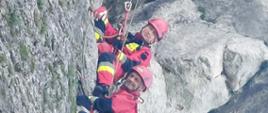 Na zdjęciu widać ratowników wysokościowych w kombinezonach wysokościowych w kolorze czerwonym. Zdjęcie wykonane w górach, w ciągu dnia. Skały w kolorze jasnoszarym, częściowo pokryte mchem.
