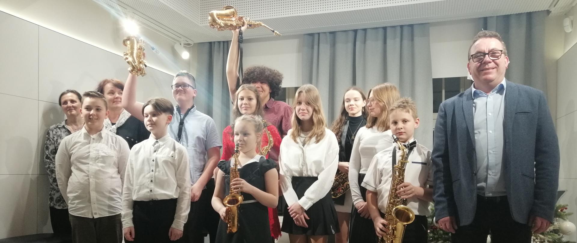 Zdjęcie przedstawia uczniów klasy saksofonu i klasy fortepianu wraz z pedagogami stojących w dwurzędzie na scenie. Niektórzy uczniowie trzymają w rękach saksofon. Dwóch chłopaków uniosło saksofony do góry. Z tyłu widać okna, kotary i ubraną choinkę. 