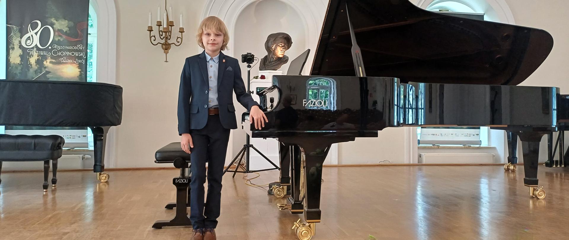 chłopiec w garniturze stojący przy fortepianie w zabytkowej białej sali, za nim baner festiwalowy i popiersie Fryderyka Chopina