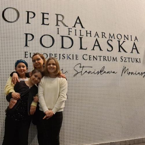 Zdjęcie przedstawia trzy uczennice i ich nauczycielkę, stojące na tle białej ściany z logo Opery i Filharmonii Podlaskiej - Europejskiego Centrum Sztuki im. S. Moniuszki.
