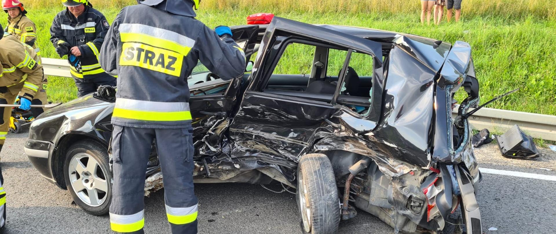 Uszkodzony samochód z wgniecionym bokiem na drodze a przy nim strażacy.