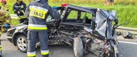 Uszkodzony samochód z wgniecionym bokiem na drodze a przy nim strażacy.
