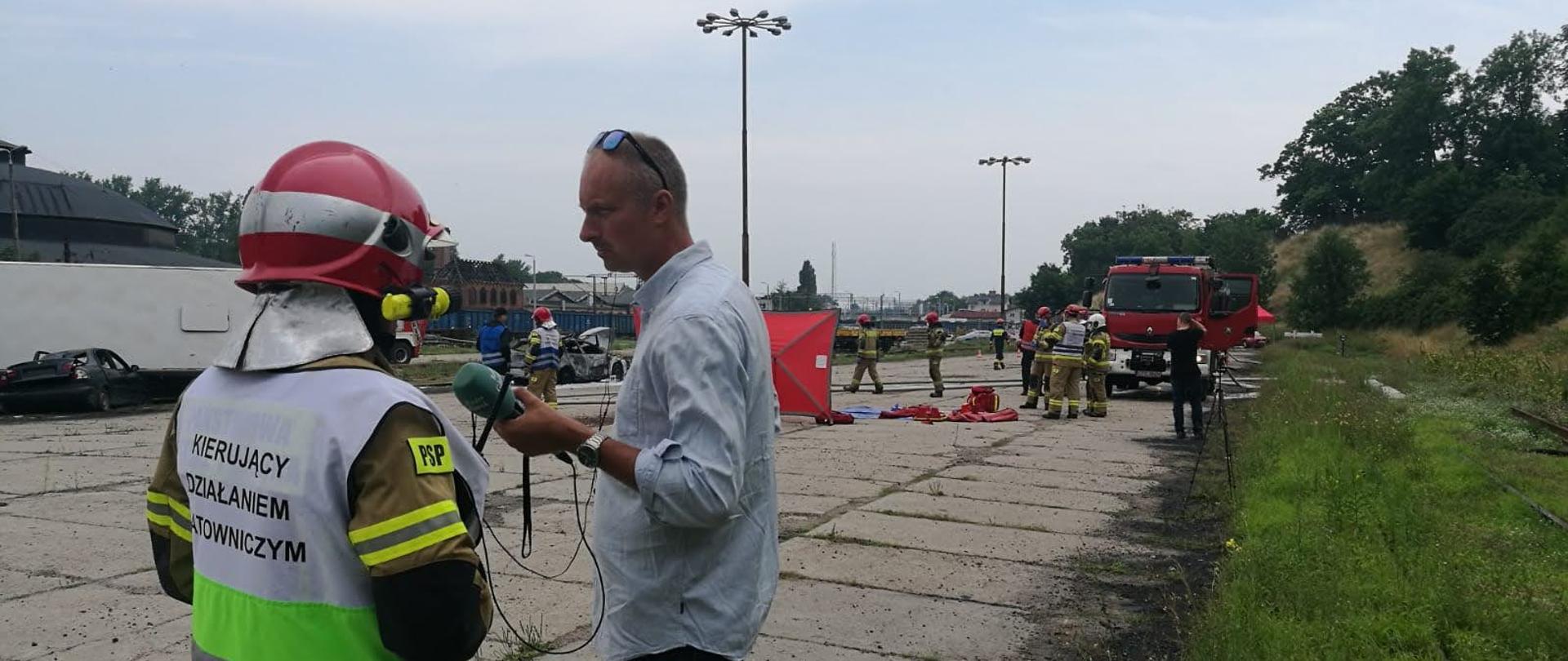 Kierujący działaniem ratowniczym udziela informacji dziennikarzowi, strażacy działają na terenie akcji.