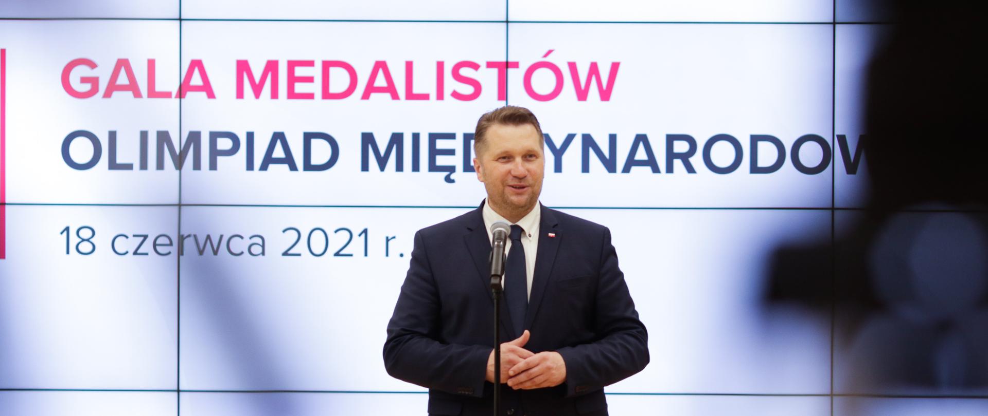 Minister Przemysław Czarnek stoi przy mikrofonie. W tle duży ekran z napisem Gala medalistów olimpiad międzynarodowych 18 czerwca 2021 r. Z prawej strony rozmazany widok kamery telewizyjnej. 