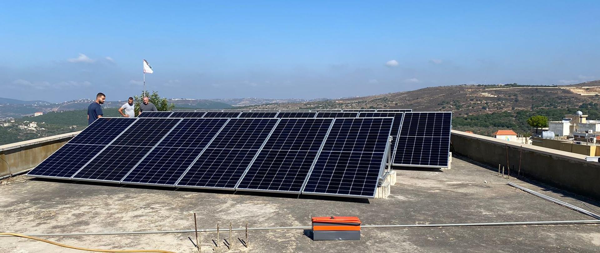 Panele fotowoltaiczne na dachu budynku, w tle widać wzgórza