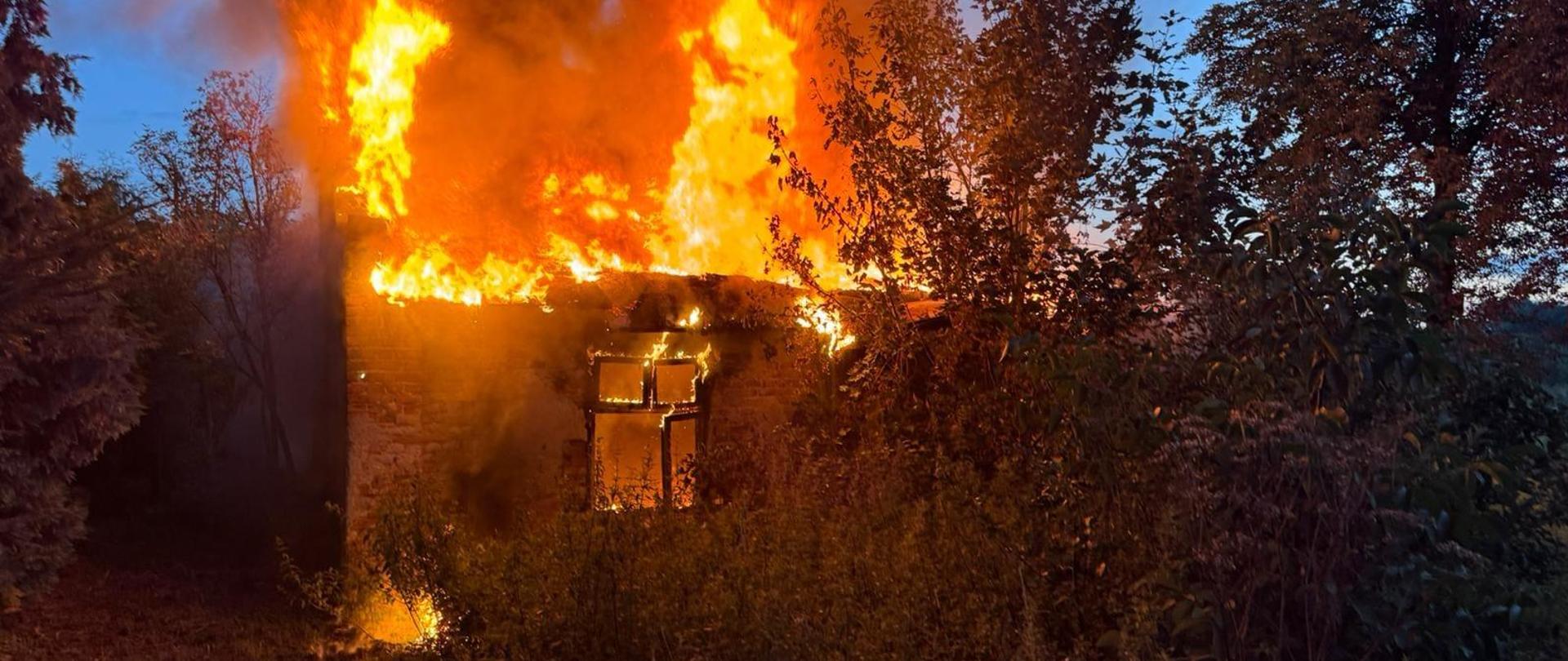 Na zdjęciu murowany budynek cały objęty ogniem. Budynek jest bez dachu. Dookoła widać krzewy. Jest półmrok.