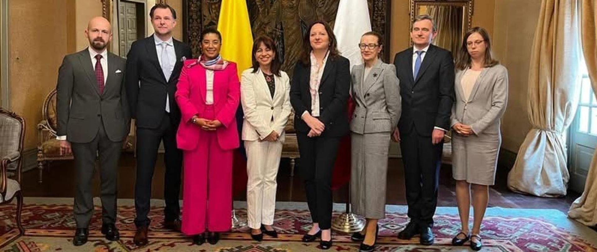 Na zdjęciu stoi osiem elegancko ubranych osób. W tle stoją dwie flagi