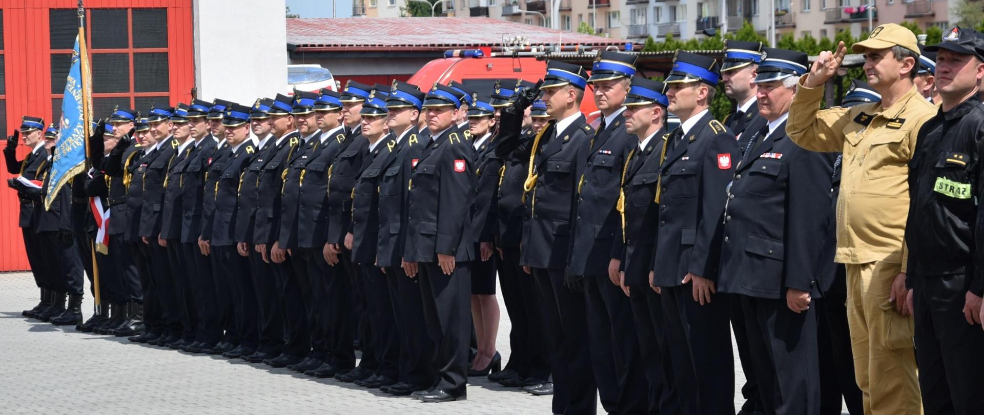 Kompania strażaków ustawiona w szeregu na placu. Dowódcy pododdziałów salutują.