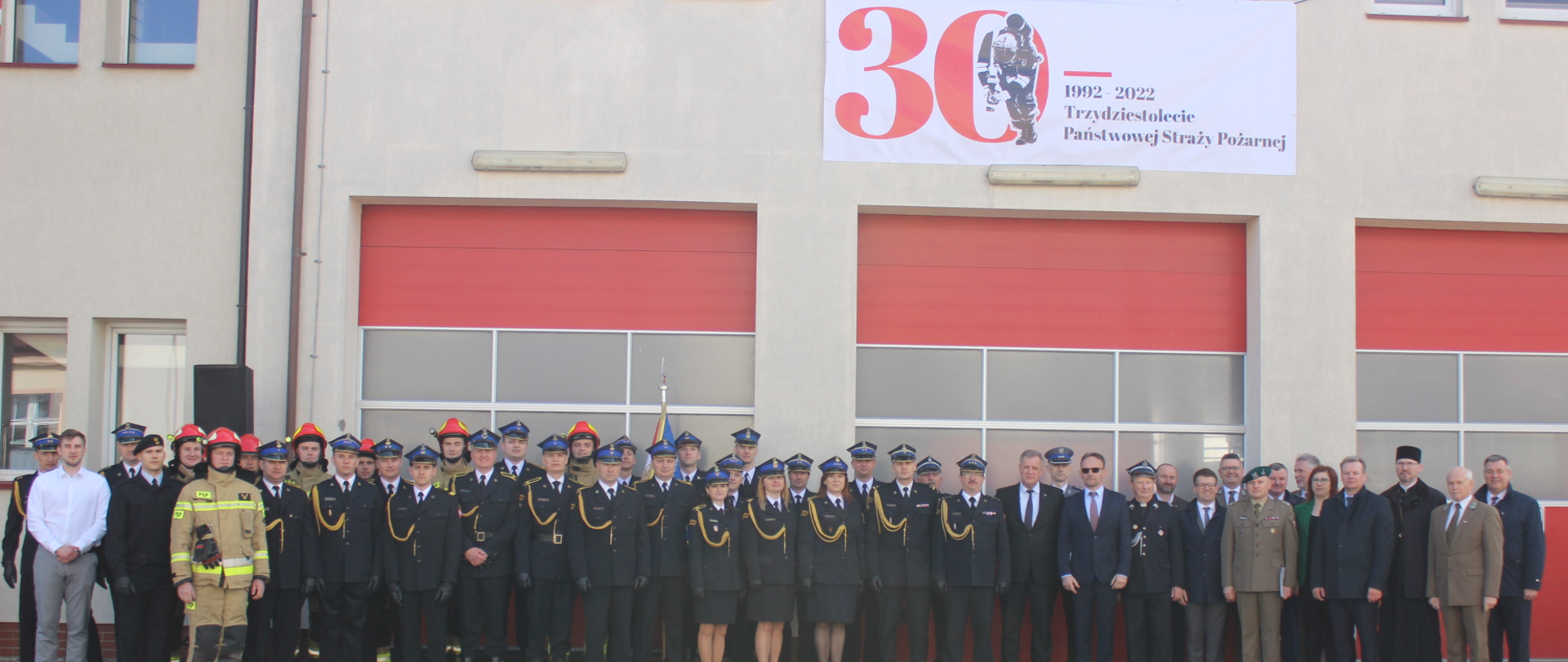 Zdjęcie grupowe podczas uroczystości powołania komendanta powiatowego, na budynku wisi biały baner 30lecie Państwowej Straży Pożarnej