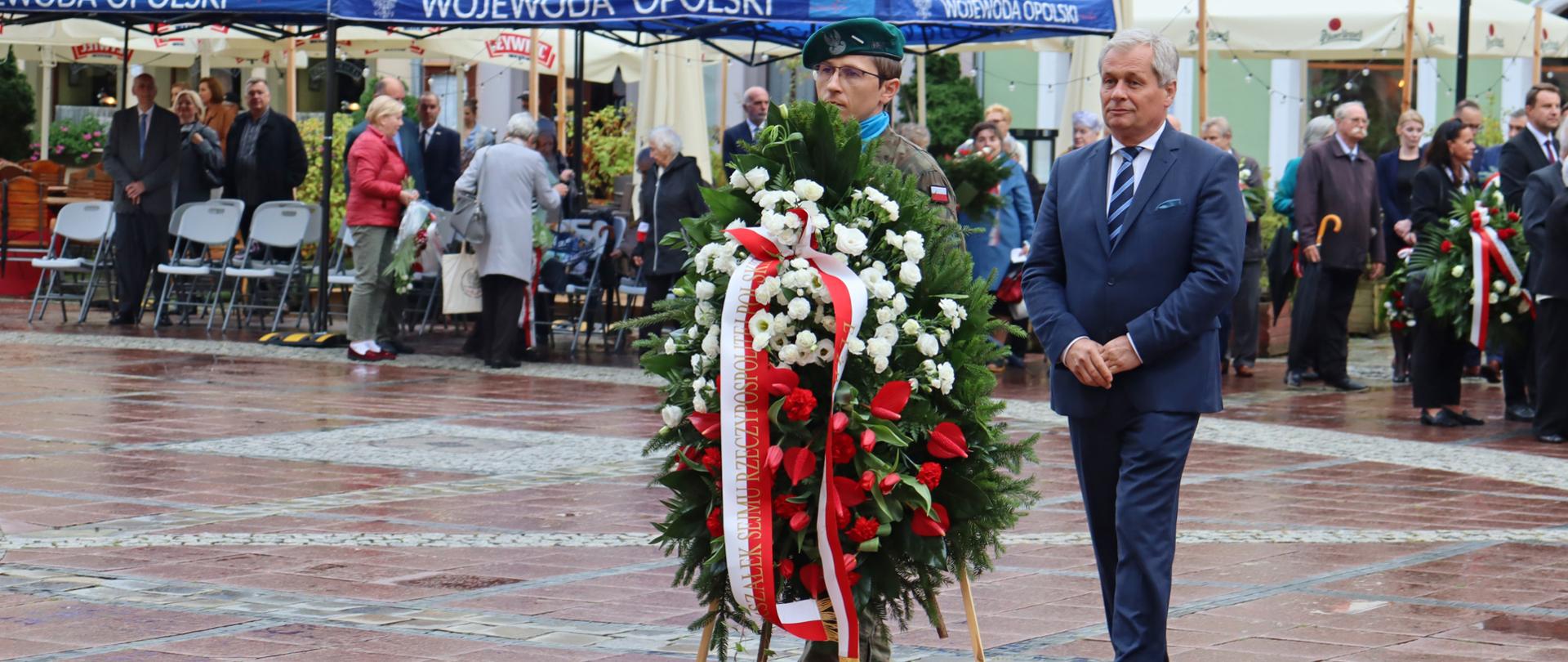 Na zdjęciu wojewoda opolski, który idzie za żołnierzem, niosącym wieniec z biało-czerwonymi kwiatami i szarfą Marszałek Sejmu RP. W tle kawiarenki, ulokowane na opolskim rynku. Po prawej stronie kadru widać granatowy namiot z napisem Wojewoda Opolski. 