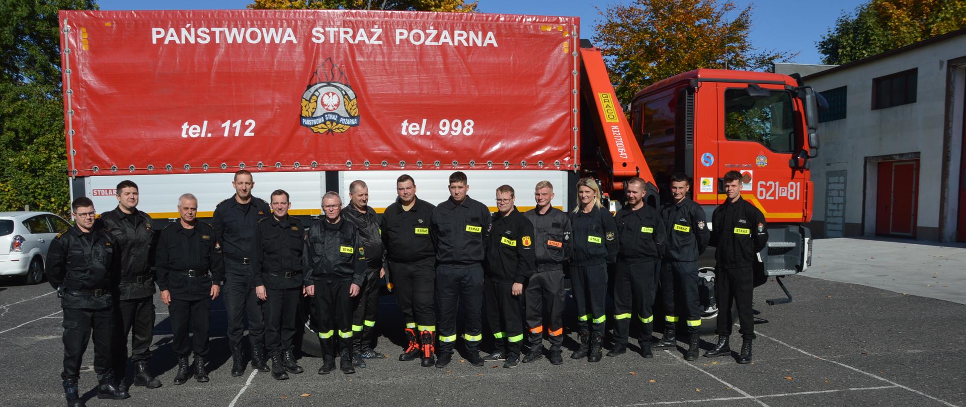 Szkolenie kierowców. Plac Komendy Powiatowej PSP w Rawiczu. Na tle samochodu kwatermistrzowskiego, który będzie wykorzystywany do zajęć praktycznych, stoją słuchacze wraz z przedstawicielami Komendy Wojewódzkiej PSP w Poznaniu oraz kierownictwem KP PSP w Rawiczu. Na czerwonej plandece samochodu Jest napis "PAŃSTWOWA STRAŻ POŻARNA", logo PSP oraz numery alarmowe. Z prawej strony widoczny jest fragment budynku, a w tle konary drzew.