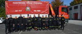 Szkolenie kierowców. Plac Komendy Powiatowej PSP w Rawiczu. Na tle samochodu kwatermistrzowskiego, który będzie wykorzystywany do zajęć praktycznych, stoją słuchacze wraz z przedstawicielami Komendy Wojewódzkiej PSP w Poznaniu oraz kierownictwem KP PSP w Rawiczu. Na czerwonej plandece samochodu Jest napis "PAŃSTWOWA STRAŻ POŻARNA", logo PSP oraz numery alarmowe. Z prawej strony widoczny jest fragment budynku, a w tle konary drzew.