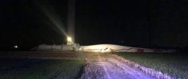 Tuż przy turbinie wiatrowej leży bardzo duży element śmigła turbiny koloru białego. Jest noc a teren jest oświetlany i zabezpieczany przez strażaków.