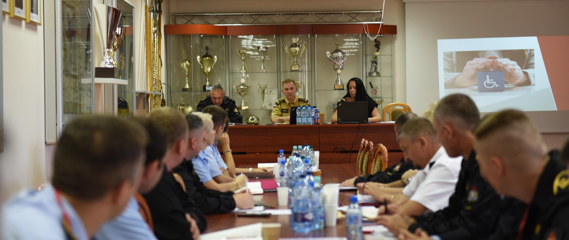 na zdjęciu 12 osób siedzących na krzesłach wzdłuż stołu w trakcie szkolenia, oraz 3 osoby przy stole na podwyższeniu za siedzącymi na ścianie widoczne są gabloty ze sztandarami i półki z pucharami