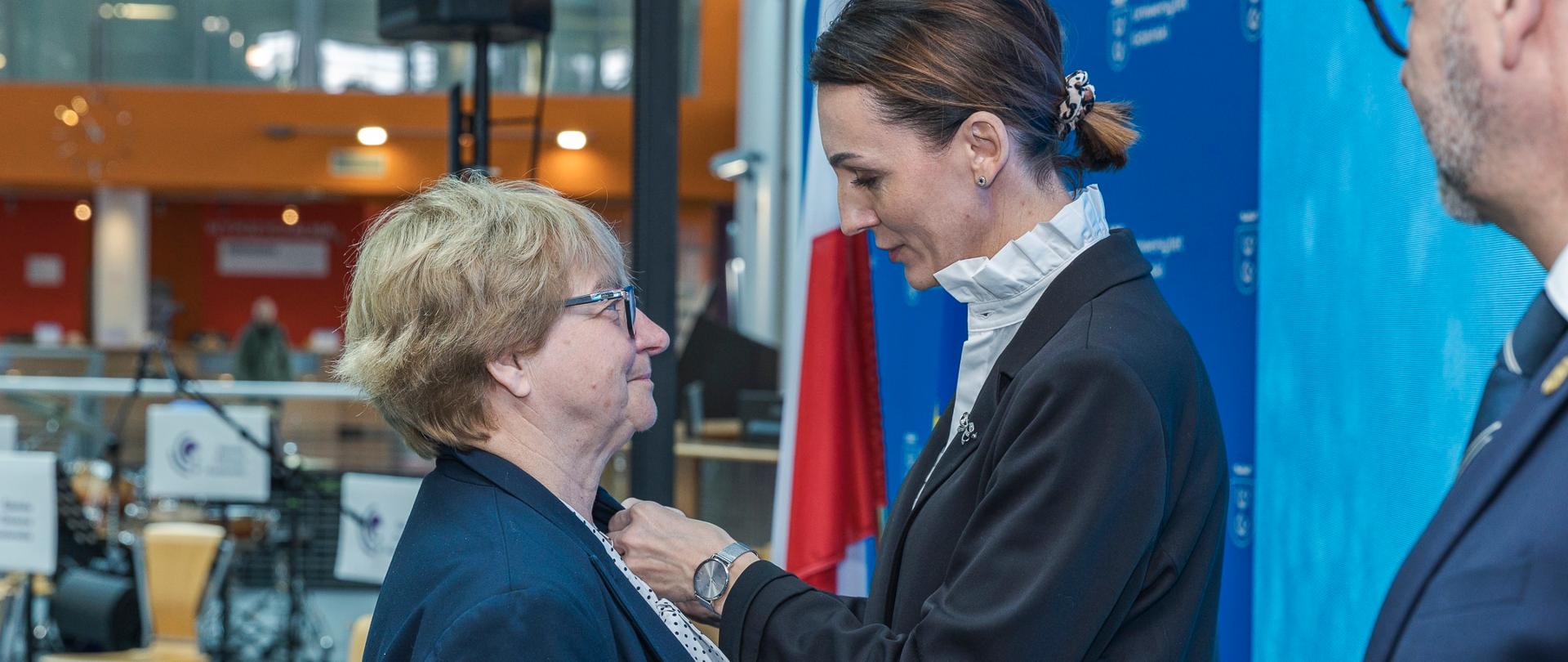 Dwie kobiety, jedna przypina medal, duża sala, flaga biało czerwona