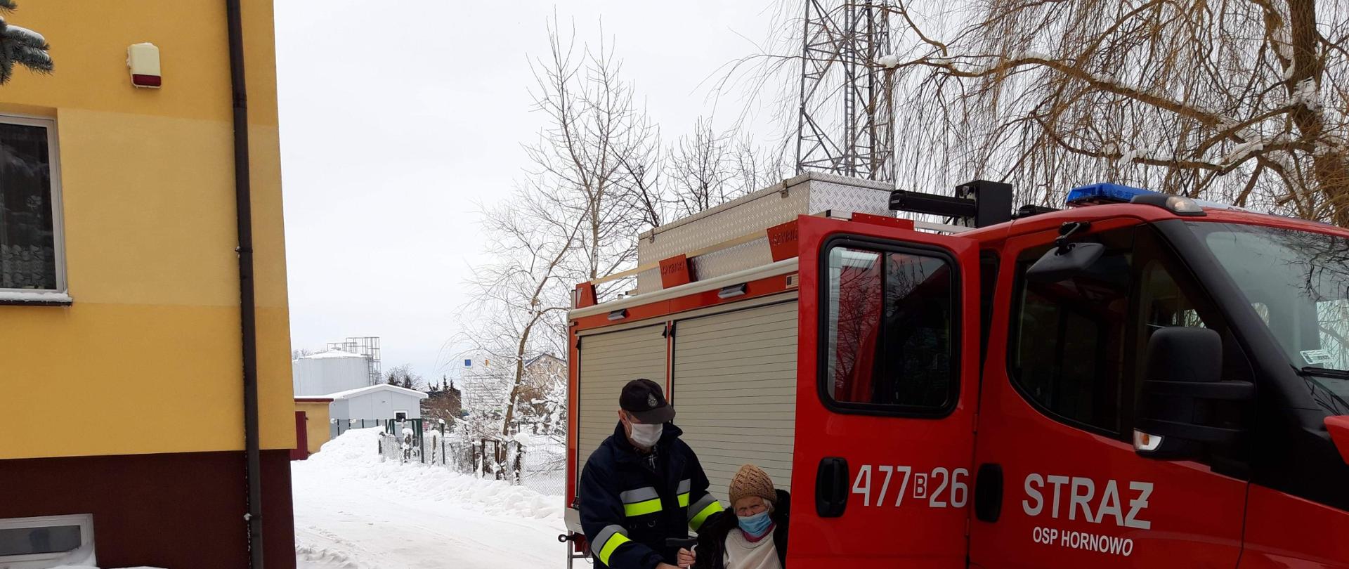 Samochód pożarniczy koloru czerwonego, stojący na śniegu. Strażak pomaga wsiadać osobie starszej która ma dojechać na szczepienie COVID-19.