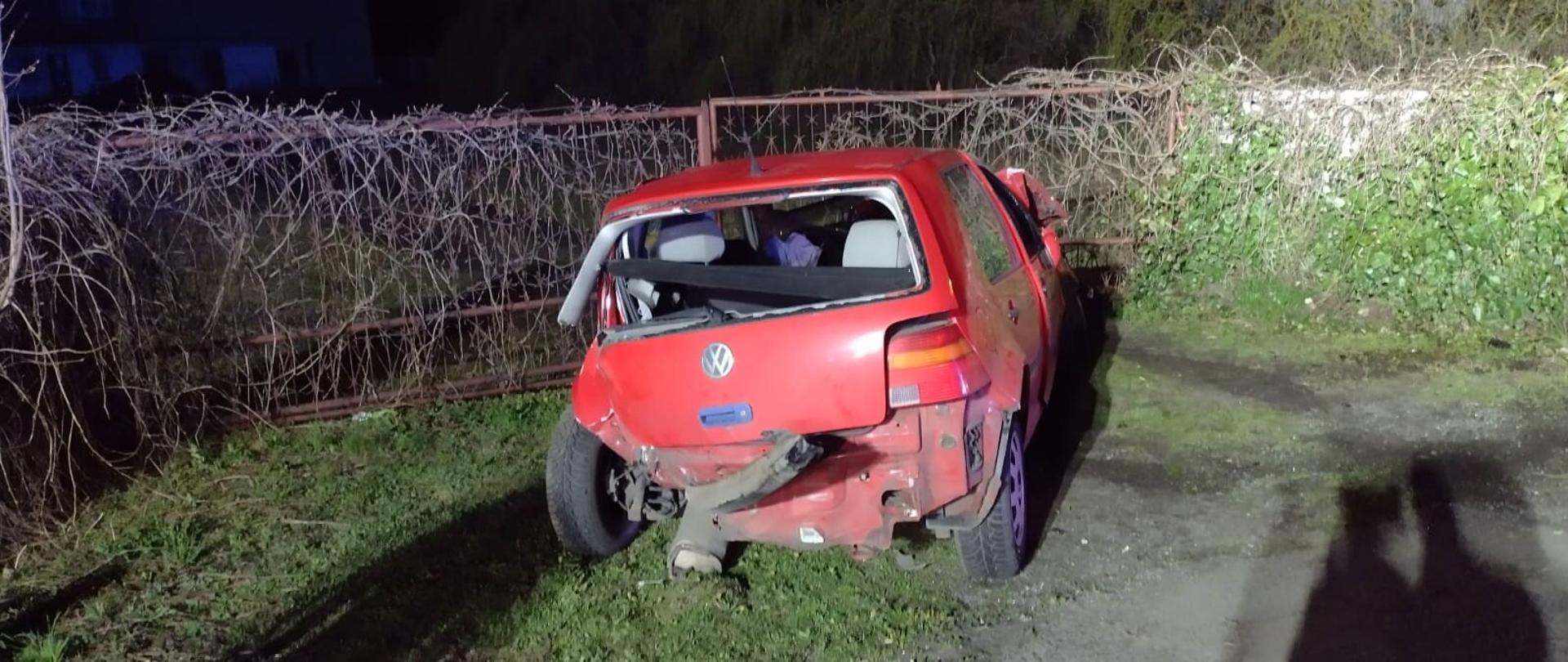 Na zdjęciu widać czerwony uszkodzony pojazd. W porze nocnej. Zdjęcie wykonane od tyłu pojazdu. Za pojazdem ogrodzenie porośnięte roślinnością. W oddali budynek mieszkalny. Teren oświetlany.