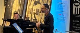 Zdjęcie przedstawia nauczyciela grającego na saksofonie z towarzyszeniem akompaniatora przy fortepianie podczas koncertu w holu Pałacu Radolińskich.