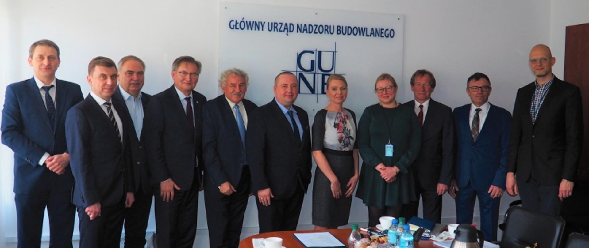 Jedenaście osób w formalnym otoczeniu biurowym, na tle napisu „Główny Urząd Nadzoru Budowlanego (GUNB)”, przed stołem z dokumentami i napojami.