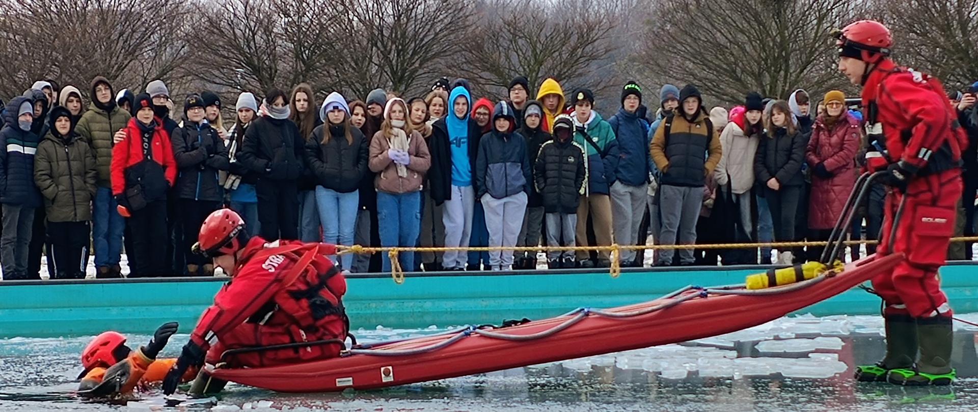 Dwóch strażaków z wykorzystaniem sań lodowych wyciąga z przerębla trzeciego strażaka