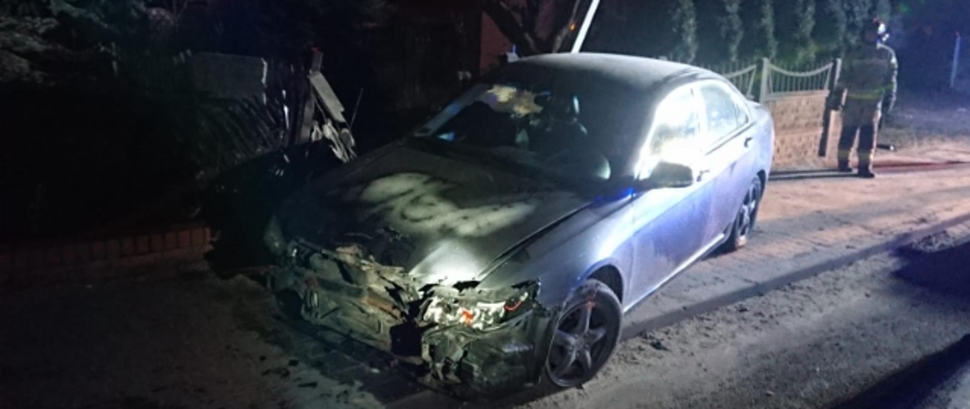 Uszkodzony przód Hondy Accord. W tle zniszczony płot i pochylona latarnia uliczna. Za pojazdem strażak zabezpieczający teren działań. 