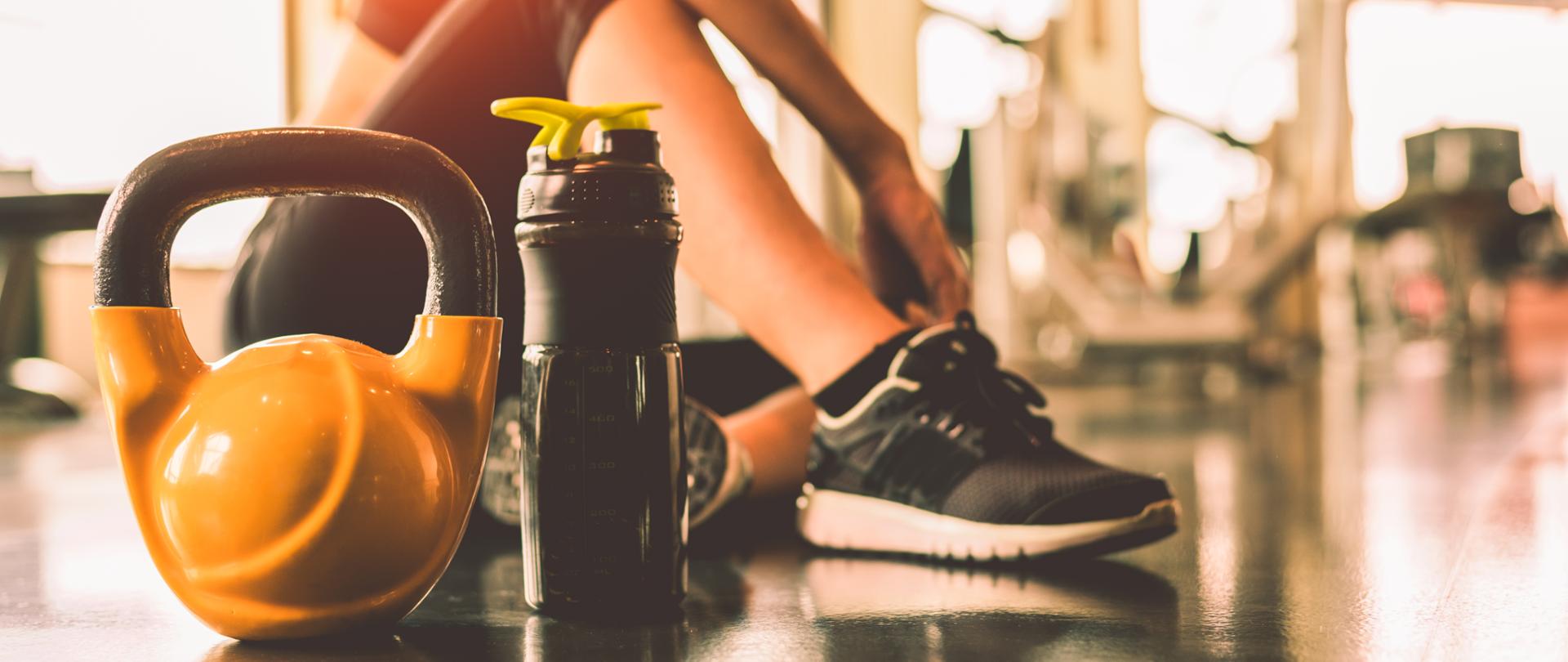 Kobieta odpoczywa po wykonanych ćwiczeniach na siłowni. Na pierwszym planie bidon oraz ciężarek.