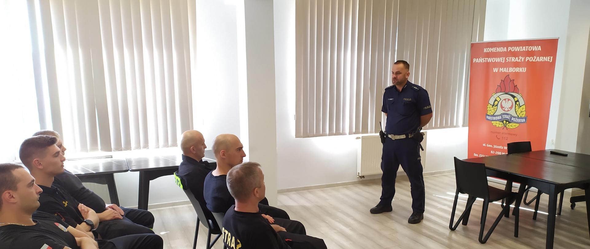 Funkcjonariusz policji stoi na przeciw siedzących strażaków i prowadzi szkolenie.