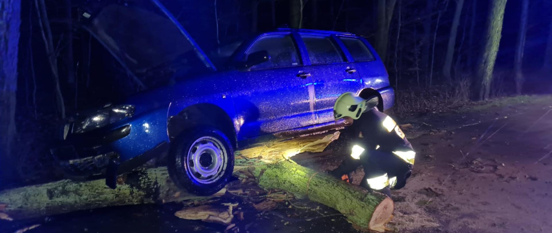 Zdjęcie wykonane w porze nocnej. Na zdjęciu widoczny samochód osobowym (kombi) koloru niebieskiego, który najechał na konar drzewa na jezdni. Obok samochodu, strażak OSP w ubraniu specjalnym koloru czarnego z białym hełmem, tnie piłą spalinową (w pozycji kucniętej) kawałek konaru drewna. W tle widoczny las.