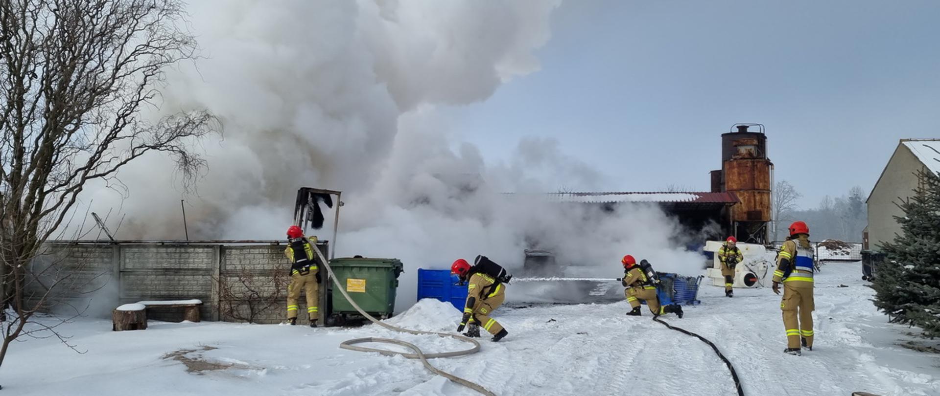 Strażacy w czasie działań ratowniczo – gaśniczych odpowiednio zabezpieczeni Aparatami ochrony dróg oddechowych walczą z pożarem, który rozwinął się na jednej z posesji. Przed funkcjonariuszami Straży Pożarnej widoczne są kłęby gęstego, białego dymu. 