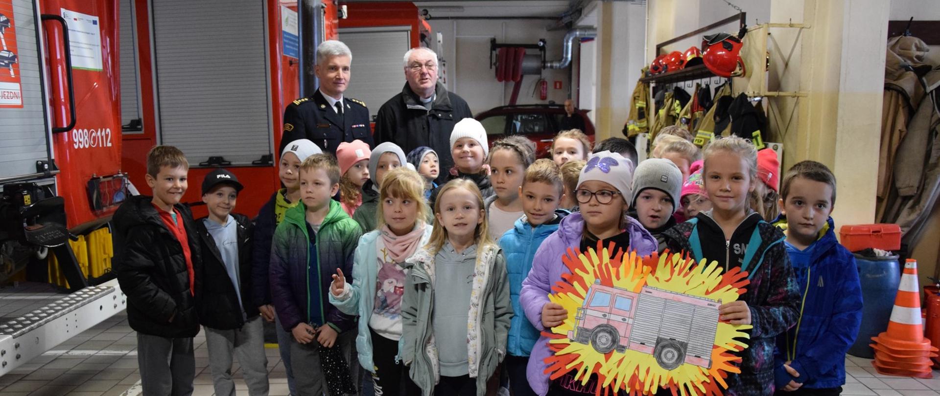 dzieci na garażu zapoznają się ze sprzętem jakim dysponują mińscy strażacy w tle komendant powiatowy PSP i kapelan strażaków