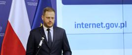 Minister cyfryzacji Janusz Cieszyński przemawia do mikrofonu, za jego plecami flagi Polski i UE oraz grafika informująca o stronie internet.gov.p
