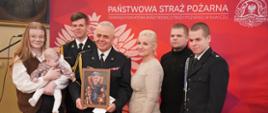 Grupa siedmiu osób stoi blisko siebie na tle czerwonej ścianki z napisem „Państwowa Straż Pożarna”. W centrum znajduje się starszy funkcjonariusz PSP w galowym, czarnym mundurze ze złotym sznurem i odznaczeniami. Trzyma w rękach oprawiony obraz przedstawiający jego portret w humorystycznej formie. Po jego bokach stoją bliscy – kobieta w jasnej sukience, młodzi mężczyźni w eleganckich strojach oraz funkcjonariusz w mundurze. Z lewej strony kobieta trzyma na rękach niemowlę. Wszyscy patrzą w stronę aparatu, a ich twarze wyrażają dumę, radość i wzruszenie, podkreślając rodzinny i uroczysty charakter chwili.