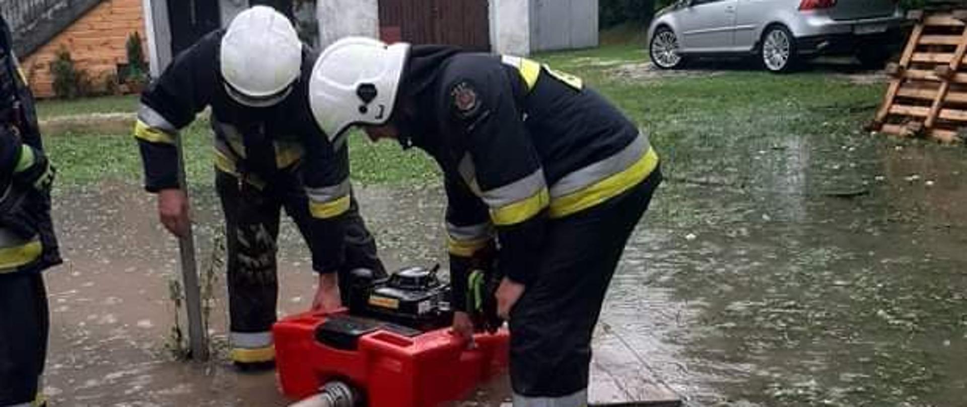 W centralnej części zdjęcia widać trzech strażaków ubranych w ubranie specjalne i białe hełmy. Strażacy stoją w niewielkim rozlewisku na posesji. Dwóch z nich obsługuję czerwoną pompę pływającą, do której podłączony jest wąż tłoczny W75. Jeden z nich się przygląda. Na drugim planie, w centralnej części widać budynek mieszkalny. W prawej części fotografii zauważyć można stojący samochód marki VW i drugi obiekt mieszkalny, znajdujący się na sąsiedniej posesji.