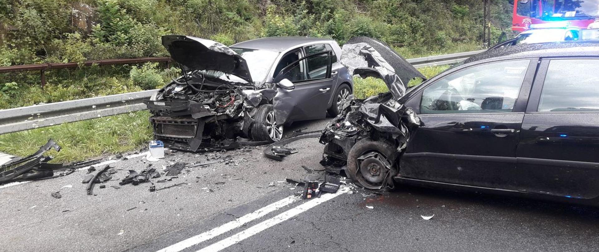 Widok przodów rozbitych samochodów. Jeden stoi w poprzek drogi a drugi na poboczu.