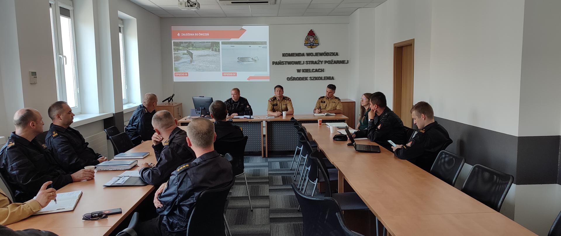 Zdjęcie przedstawia oficerów Państwowej Straży Pożarnej - za nimi ekran z prezentacją dot. omawianych ćwiczeń oraz stół prezydialny z kierownictwem KW PSP i KM PSP Kielce