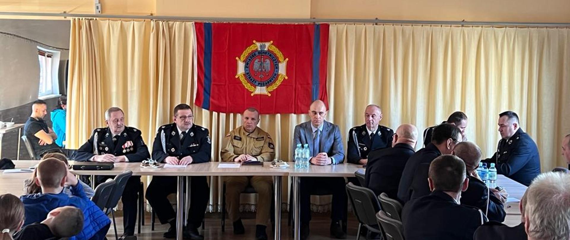 Zdjęcie zrobione na świetlicy. Trwa zebranie. Za stołami na wprost, z prawej i lewej strony siedzą strażacy w mundurach. Na ścianie wisi na czerwonym tle logo związku ochotniczych straży pożarnych.